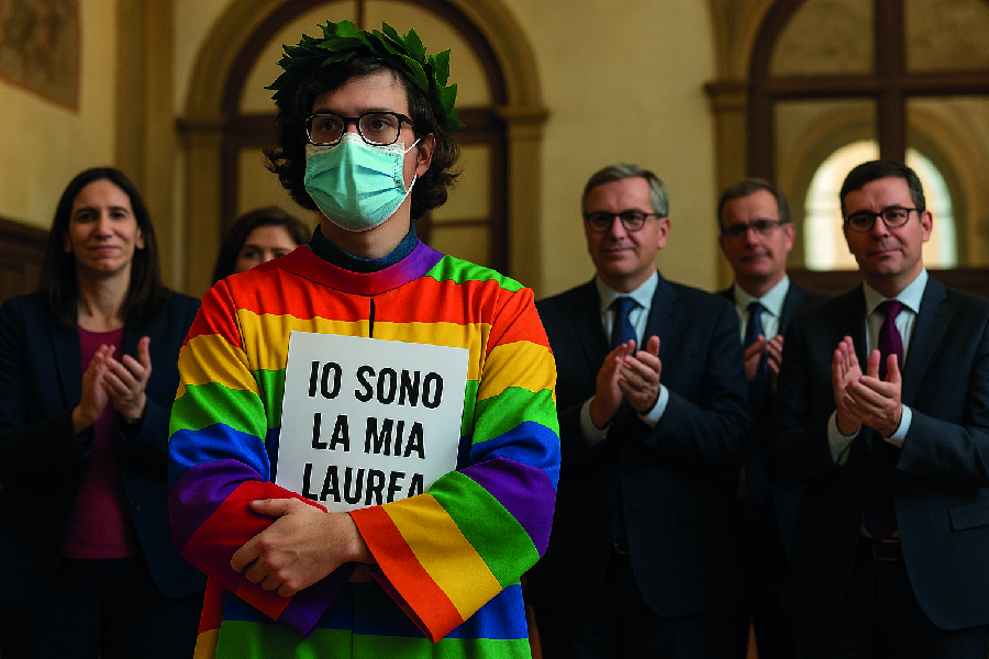 Studente non-binario con cartello “IO SONO LA MIA LAUREA” durante protesta all’Università di Bologna, indossa camice arcobaleno. Schlein e Boldrini applaudono.